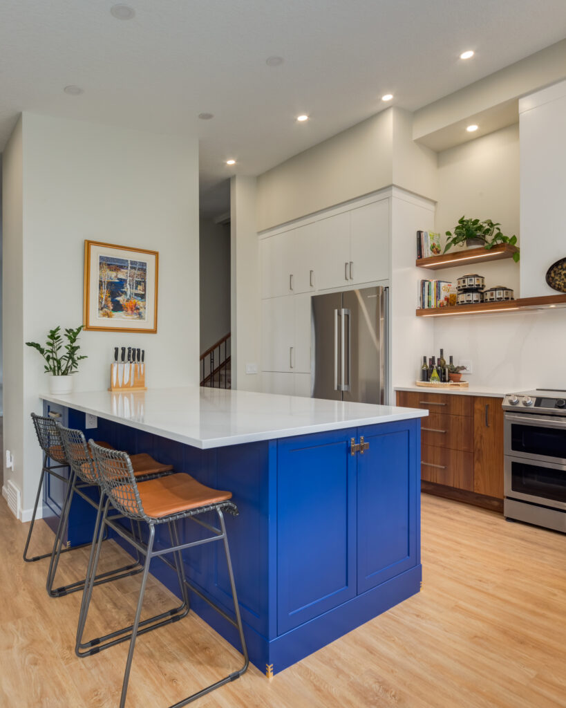 Modern kitchen with a bold blue island, two brown leather barstools, white countertops, stainless steel appliances, wood cabinets, and wall art above a plant on the left wall. Warm wood floors and recessed lighting throughout.