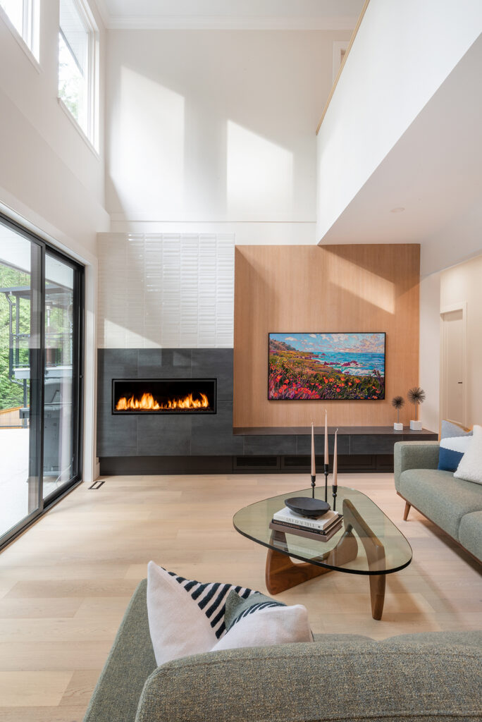 Modern living room with high ceilings, large windows, a sleek black fireplace, wall-mounted TV displaying colorful art, light wood floors, green sofas, a glass coffee table, and sliding doors opening to a patio.