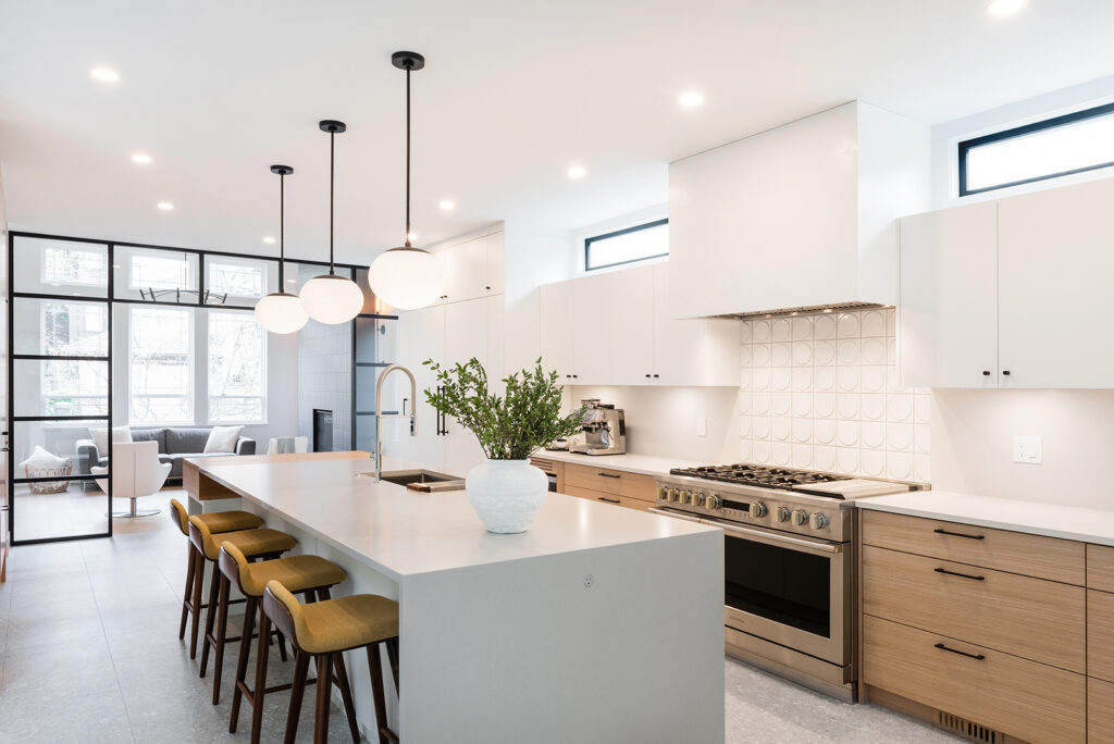 Modern kitchen with white countertops, wood cabinets, a large island with four yellow stools, pendant lights, stainless steel appliances, and a white vase with greenery. Bright and open with a view into a living area.