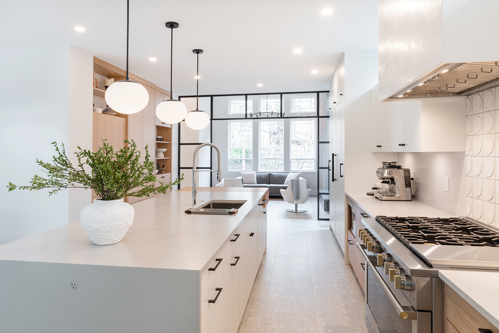 Bright, modern kitchen with white cabinetry, island with sink, hanging globe lights, stainless steel stove, built-in shelves, and a potted plant. Large windows and a living area are visible in the background.