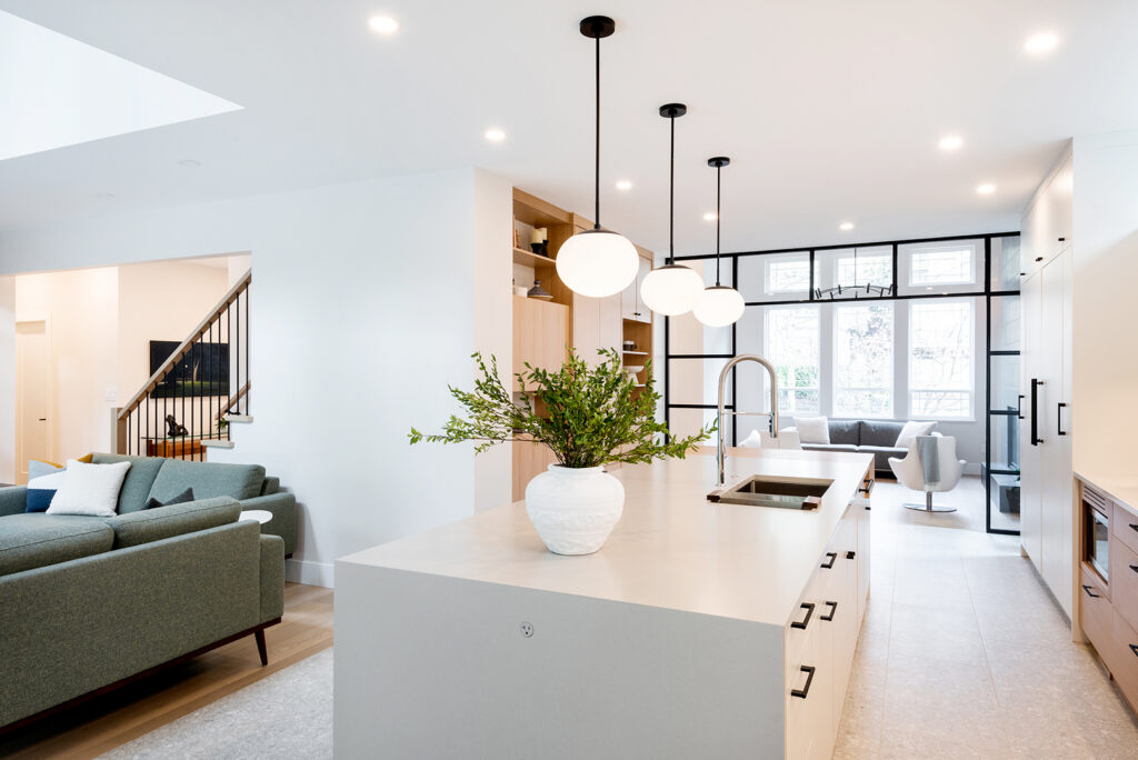 A modern, bright kitchen with white walls, a large island featuring a white countertop and a vase of greenery, pendant lights, and an open living area with a green sofa and stairs in the background.