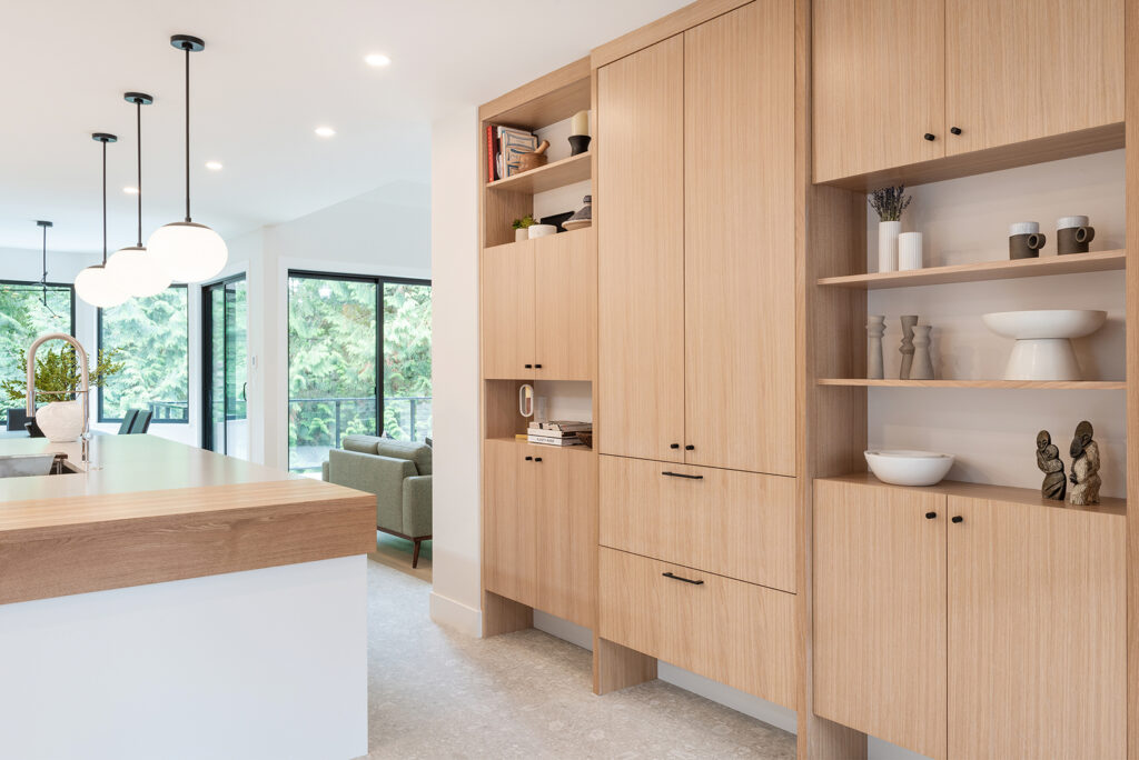 Modern kitchen with light wood cabinets, open shelves displaying decor, a white island with a wood accent, and globe pendant lights. Large windows in the background reveal greenery outside.
