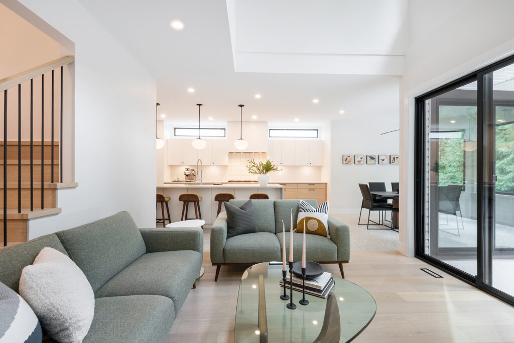 Modern open-concept living area with green sofas, a glass coffee table, and candles, adjacent to a sleek white kitchen with pendant lights and barstools, and a dining area near large sliding glass doors.