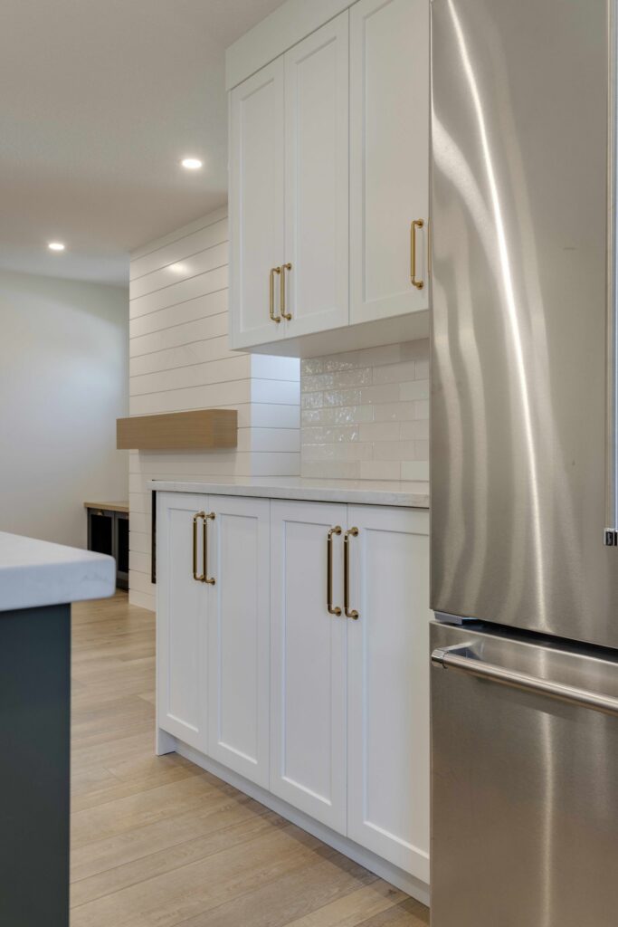 Modern kitchen with white cabinets, gold handles, light wood flooring, a stainless steel refrigerator, and white subway tile backsplash. The space is bright and clean with minimalist decor.