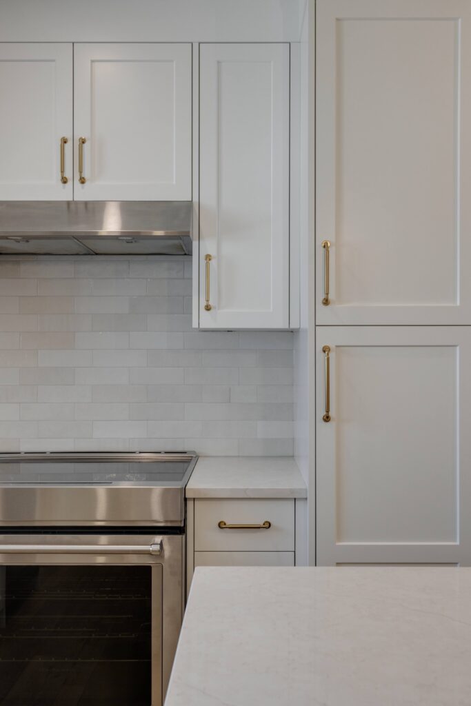 A modern kitchen with white cabinets, gold handles, a stainless steel oven, and a light subway tile backsplash. The countertop is white, and the overall look is clean and minimalistic.
