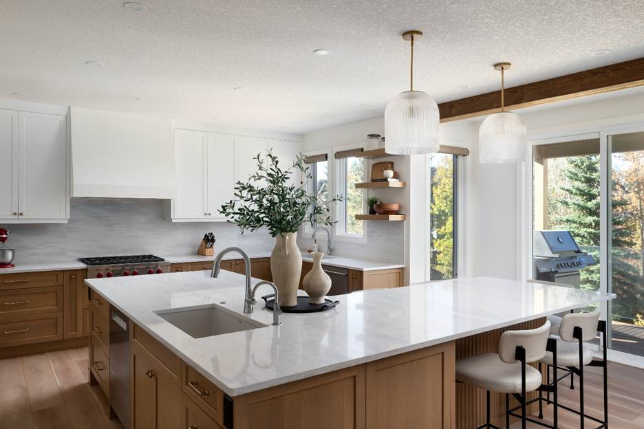 Modern kitchen with light wood cabinets, white countertops, a large island with a sink, two pendant lights, bar stools, and large windows overlooking trees. A vase with greenery sits on the island.