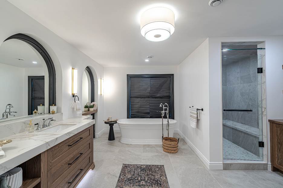 Modern bathroom with a double-sink vanity, marble countertop, large mirrors, a freestanding bathtub, walk-in glass shower, tiled floor, a basket, and a rug, all lit by a ceiling light.