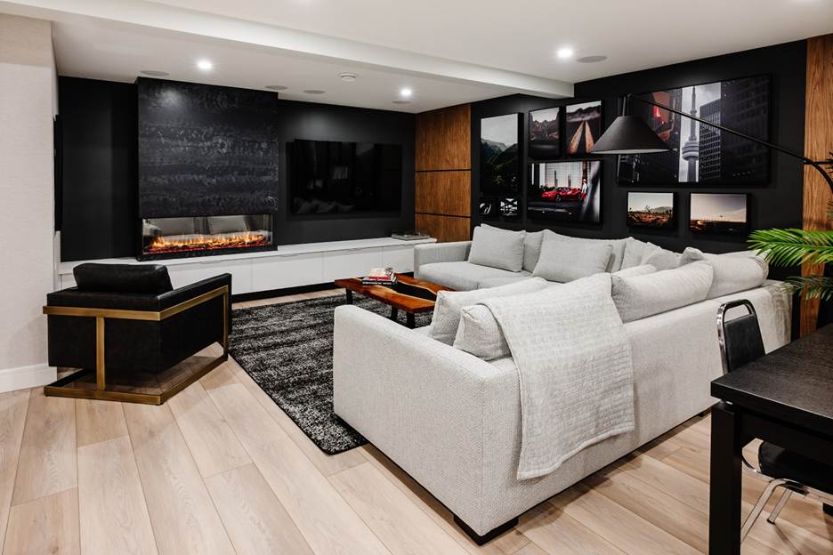 Modern living room with a large light gray sectional sofa, black armchair, wall-mounted TV, electric fireplace, wall art, black area rug, wood-paneled walls, light wood flooring, and a small table with a plant.