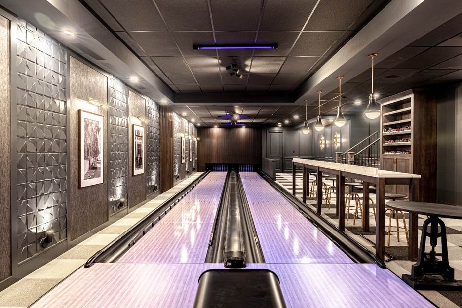 A modern, upscale home bowling alley with two lanes, stylish lighting, bar seating, wall art, and decorative wall panels in a sleek, contemporary setting.