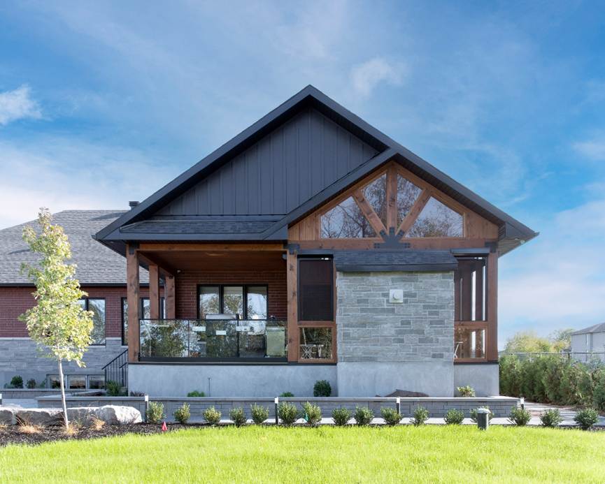 Modern single-story house with a dark gabled roof, wood and stone accents, large windows, a covered front porch, landscaped lawn, and a small tree in the front yard under a blue sky.