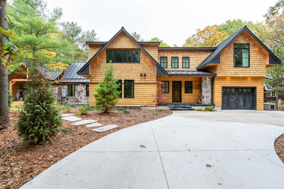 A modern two-story house with wooden siding, large windows, and a black roof sits surrounded by trees. A curved concrete driveway leads to a garage on the right, with landscaped plants along the edges.