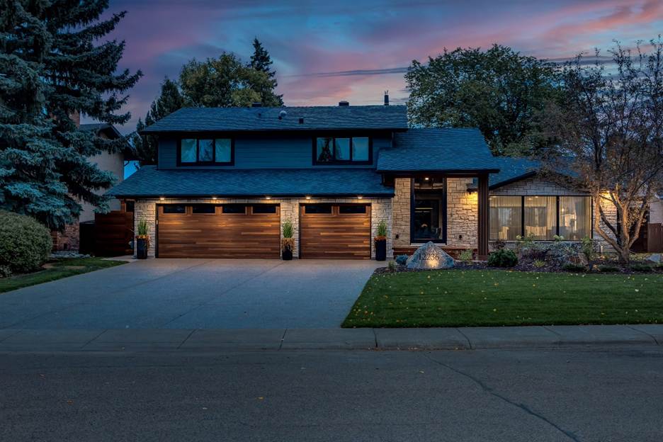 A modern two-story house at dusk with large windows, three wooden garage doors, exterior lighting, a wide driveway, green lawn, and landscaped front yard with trees and shrubs.
