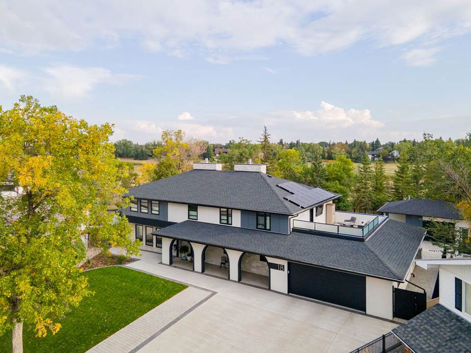 A large modern two-story house with a dark roof, light exterior walls, arched entryway, attached garage, and spacious driveway, surrounded by trees and greenery under a partly cloudy sky.