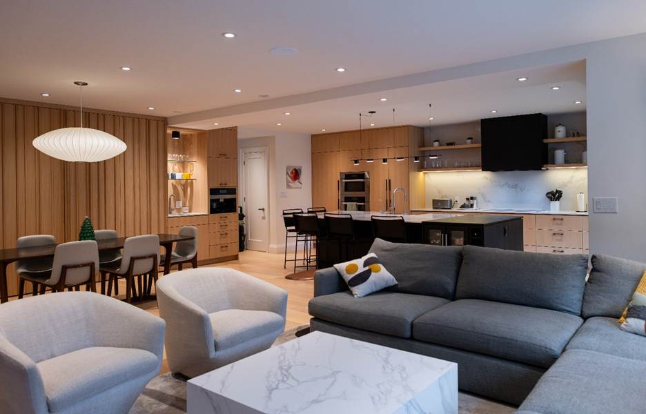 Modern open-concept living room and kitchen with light wood cabinets, a marble coffee table, gray sectional sofa, two beige armchairs, and a dining table with six chairs under a white pendant light.