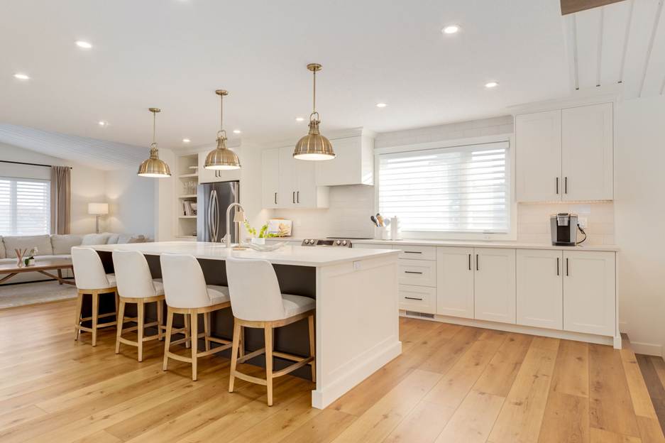 A modern kitchen with white cabinets, a large island with four white chairs, pendant lights above the island, wood flooring, and a window with blinds letting in natural light.