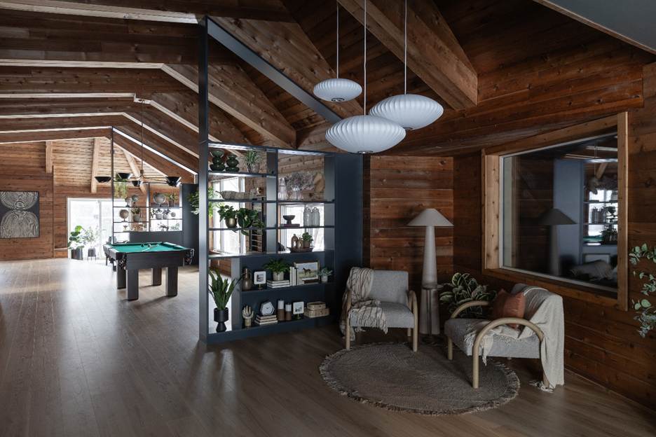 A cozy, modern living space with wooden walls and ceiling, featuring chairs, a floor lamp, indoor plants, shelves with decor, and a pool table in the background. Large windows let in natural light.