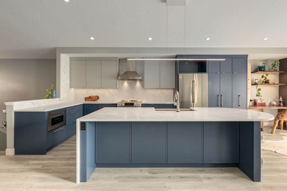 Modern kitchen with blue and white cabinets, marble countertops, stainless steel appliances, a built-in oven, and a large island with a sink. Light wood flooring and open shelves are visible in the background.