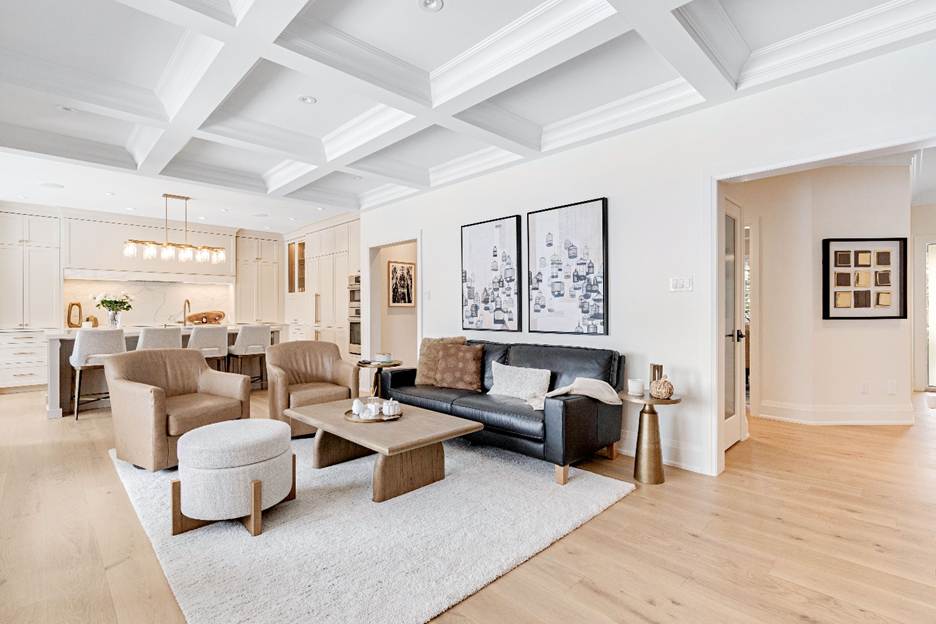 A modern open-concept living room and kitchen with light wood floors, a black sofa, two beige armchairs, a white rug, large wall art, and a white coffered ceiling. The kitchen has white cabinets and pendant lights.