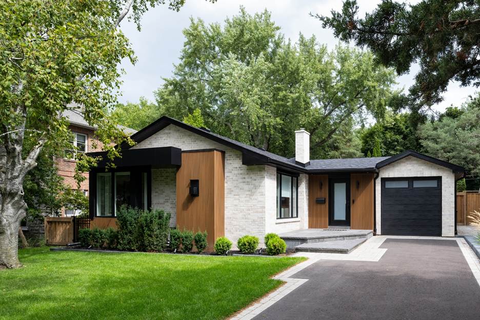 Single-story modern house with light brick and vertical wood siding, black trim, a black garage door, neatly trimmed lawn, shrubs, trees, and a paved driveway under a partly cloudy sky.