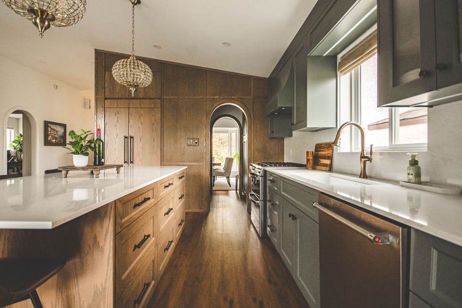 Modern kitchen with wood cabinets, a white island countertop, gray lower cabinets, stainless steel appliances, a gold faucet, pendant lights, and an arched doorway leading to a sunlit room with a chair.