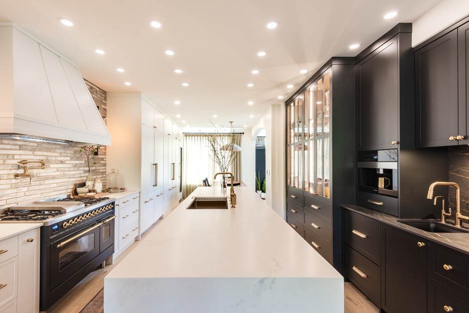 Modern kitchen with a large white island, black and white cabinetry, a built-in oven, stove, sink, and bright recessed lighting. Glass cabinets and a coffee machine are visible along the right side.