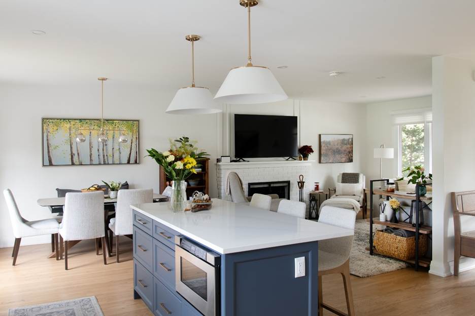 A modern open-concept living space features a kitchen island with a microwave, pendant lights, a cozy living area with a TV above a fireplace, a dining table, and large windows letting in natural light.