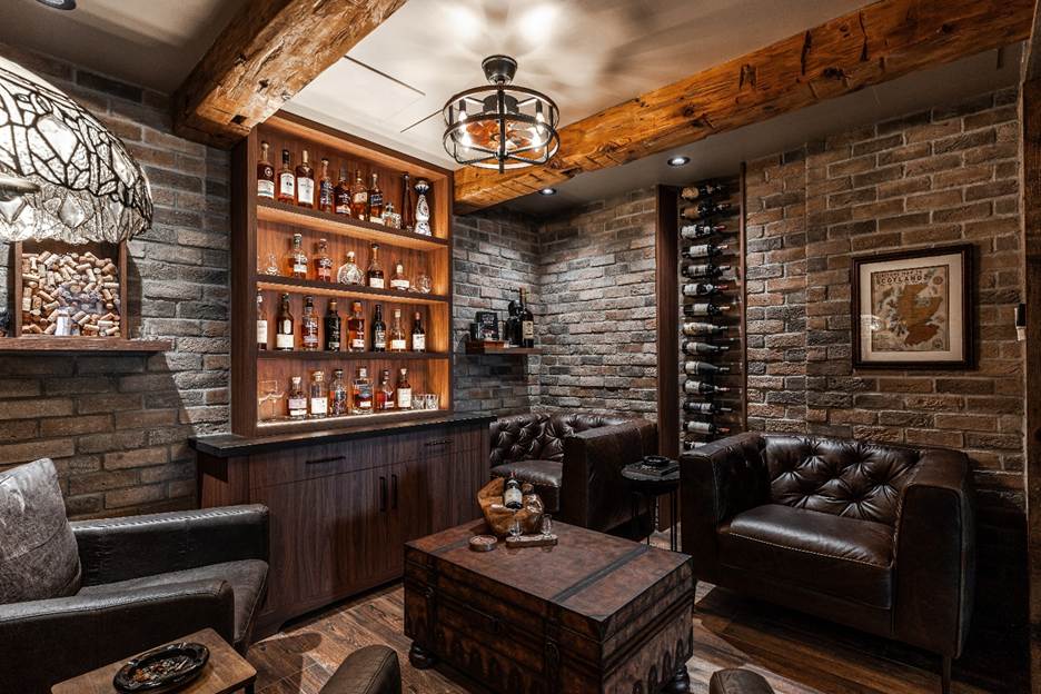A cozy, rustic lounge with exposed brick walls, leather armchairs, a wooden chest as a table, a built-in liquor shelf, a vertical wine rack, and warm lighting creating a welcoming atmosphere.