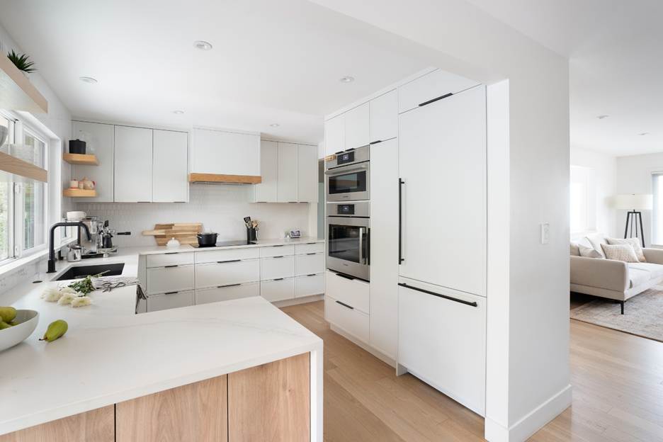 Modern, minimalist kitchen with white cabinets, built-in appliances, light wood accents, and a white countertop. Large windows provide natural light. A living room with a beige sofa is visible in the background.