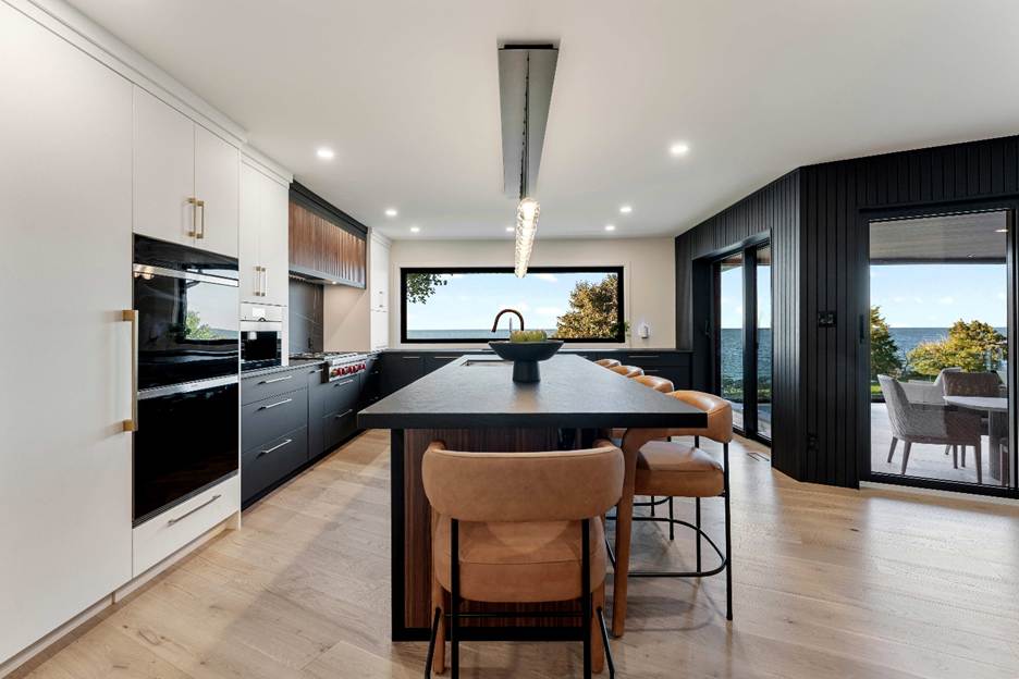 Modern kitchen with black and white cabinets, island with tan bar stools, wood flooring, large windows, and a view of greenery and water outside. Ample natural light fills the spacious room.