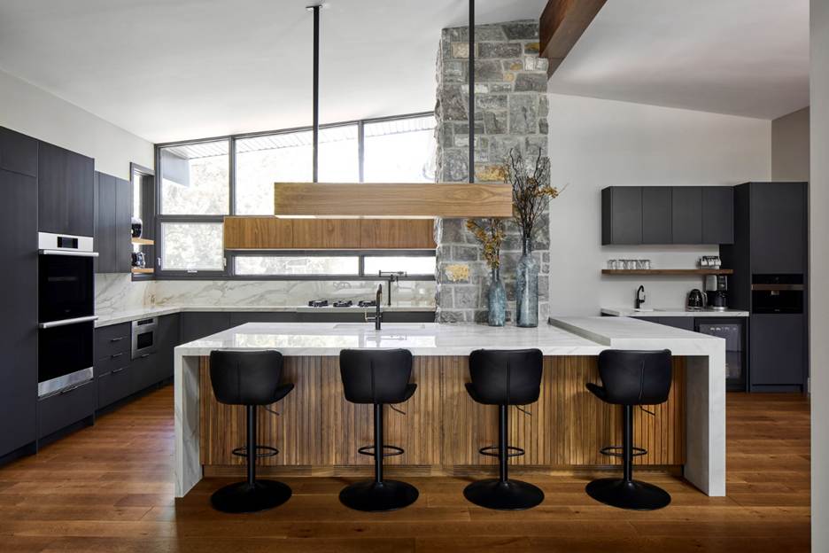Modern kitchen with a large island featuring a white countertop, four black bar stools, dark cabinets, wooden accents, a stone column, and large windows letting in natural light.