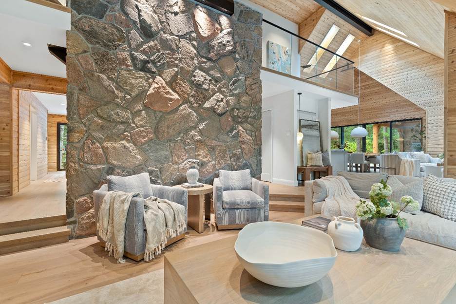 A modern living room features a large stone accent wall, cozy armchairs with blankets, wooden floors, neutral décor, and an open layout with a view into the dining area and upstairs loft.