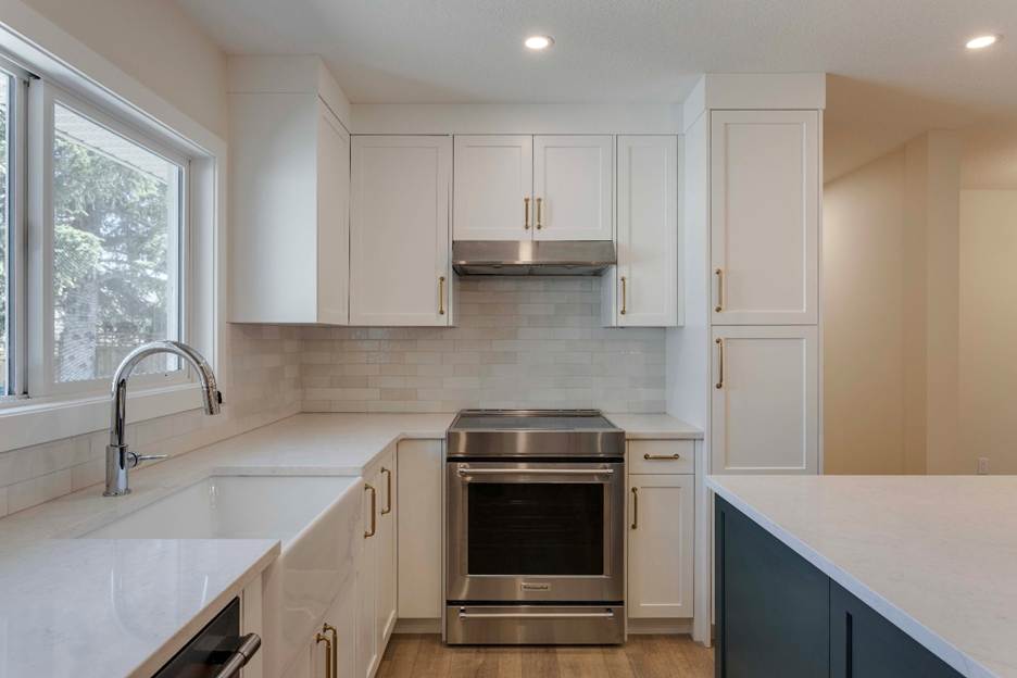 Modern kitchen with white cabinets, stainless steel oven and range hood, white subway tile backsplash, gold hardware, quartz countertops, and a large window above the sink allowing natural light to enter.