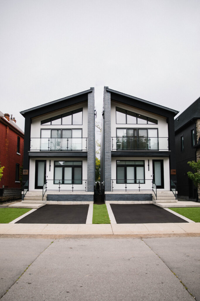 A symmetrical duplex with modern design, large windows, black trim, and glass balconies. Each unit has a staircase, driveway, and green lawn, separated by a dividing wall.