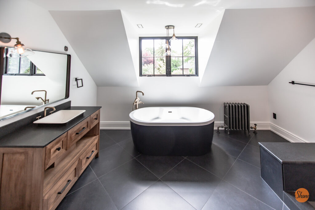 Modern bathroom with a freestanding oval tub under a window, wooden vanity with sink and mirror on the left, black tile floor, and a radiator near the window.
