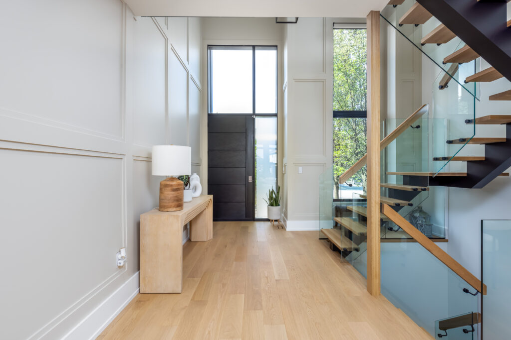 Modern entryway with light wood floors, a wooden console table with a white lamp and decor, large black front door, tall windows, and a floating staircase with glass railing. Natural light fills the space.