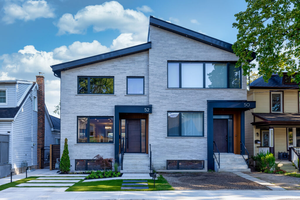 Modern duplex with light gray brick exterior, two separate entrances, black trim, large windows, small front yards, and a tree on the right. House numbers 52 and 50 are visible on the doors. Blue sky with clouds above.