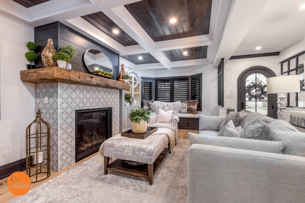 Elegant living room with a gray sofa, armchair, patterned fireplace, wooden mantel, decorative accents, plants, ottoman, and coffered ceiling with dark wood panels. Large windows and soft lighting enhance the cozy atmosphere.