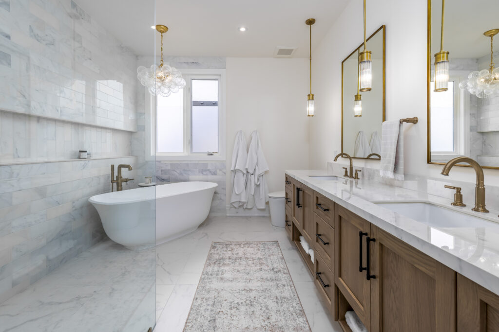 Modern bathroom with a freestanding tub, double sinks with wooden cabinets, hanging pendant lights, marble flooring and walls, a rug, and white towels hanging near a window.
