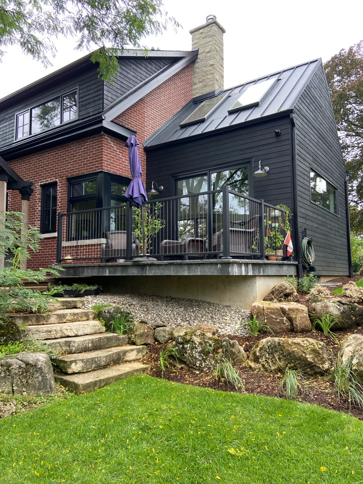 Modern house with black siding and red brick features a raised deck with outdoor furniture, railing, and a closed purple umbrella. Stone steps lead up from a landscaped yard with rocks and green grass.