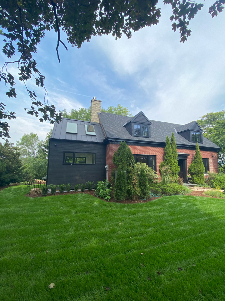 A brick house with black siding accents and dormer windows sits on a lush green lawn, surrounded by trees and shrubs under a partly cloudy sky.