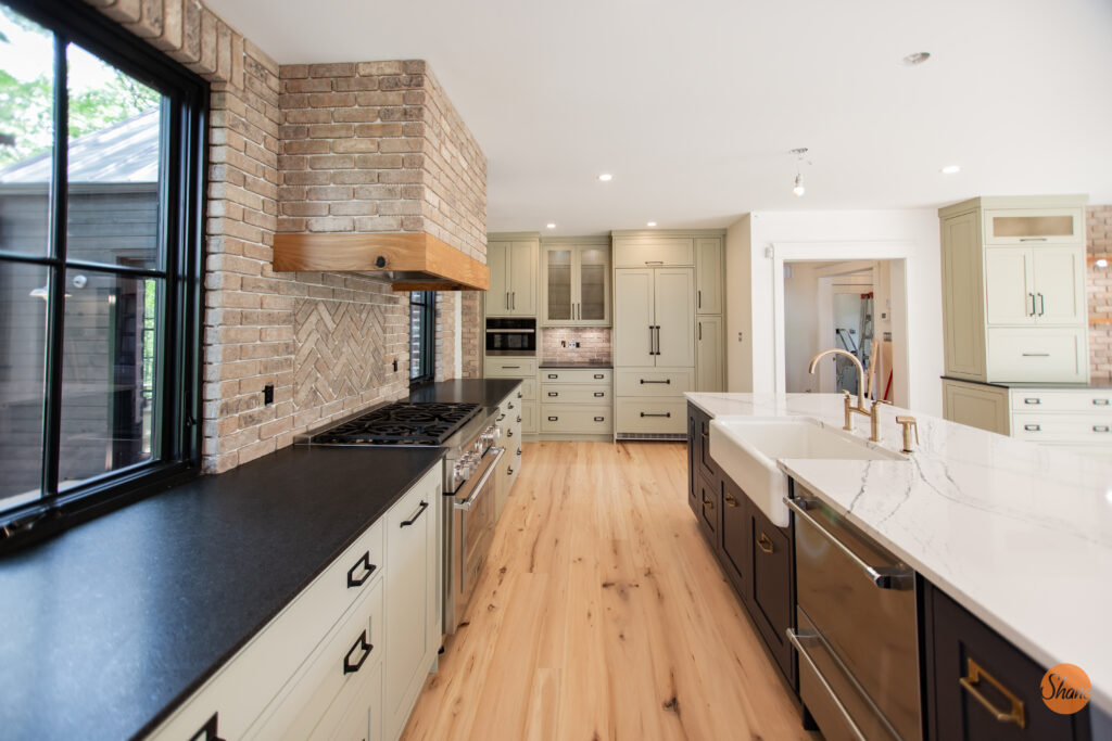 Modern kitchen with light wood floors, light green cabinets, black countertops, brick backsplash, stainless steel appliances, and a large island with a white marble surface and two sinks.