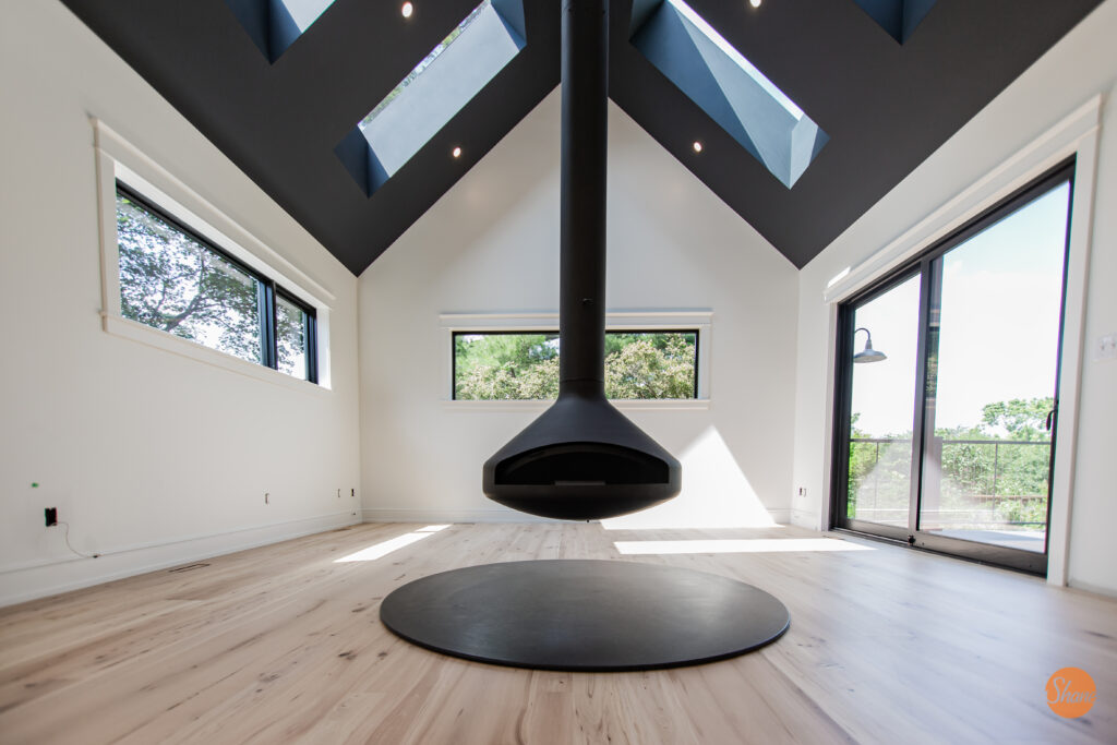 Modern, minimal living room with high ceilings, large windows, skylights, and a central black suspended fireplace over a circular mat. Light wood floors and white walls create a bright, airy space.