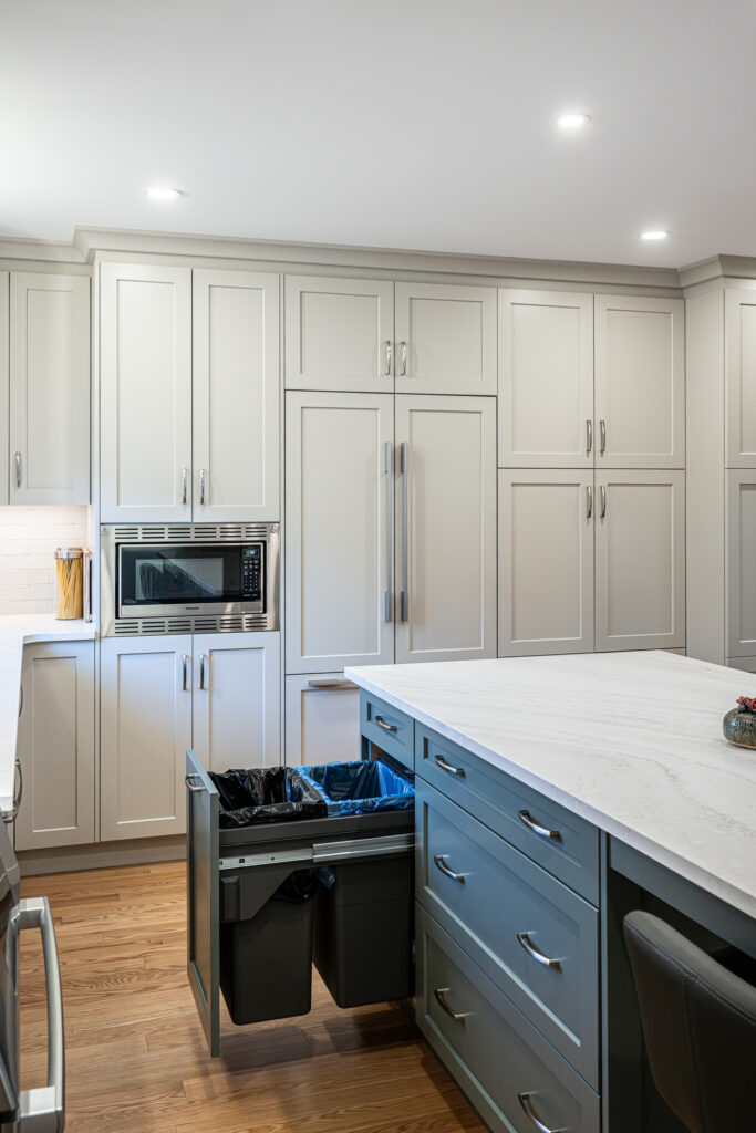 Modern kitchen with white cabinets, built-in microwave, and a large island. The island drawer is open, revealing divided pull-out trash and recycling bins. Countertops are white, and floors are wood.