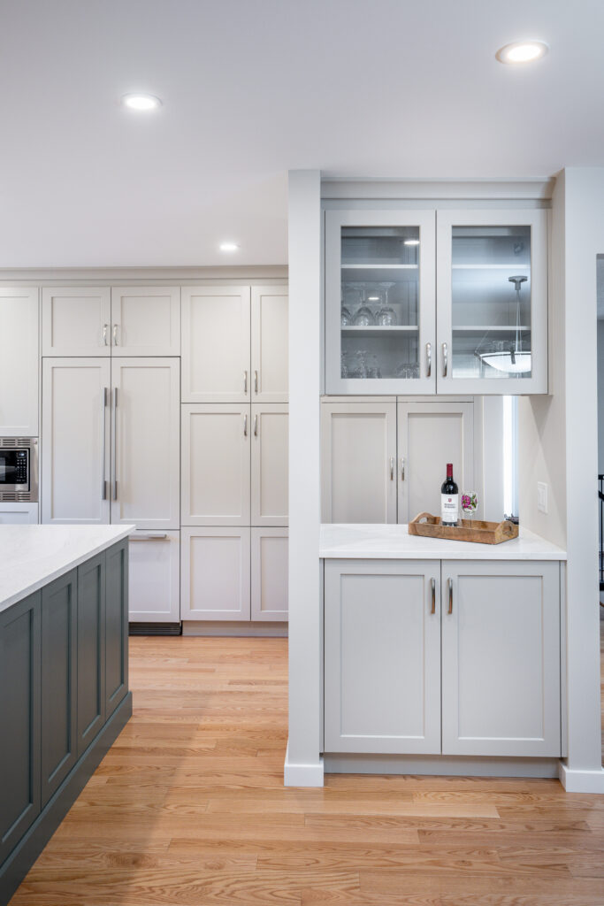 Modern kitchen with light gray cabinets, glass-front uppers, wood flooring, and a white island. A wine bottle and glass sit on a tray on the countertop beneath the upper cabinets.