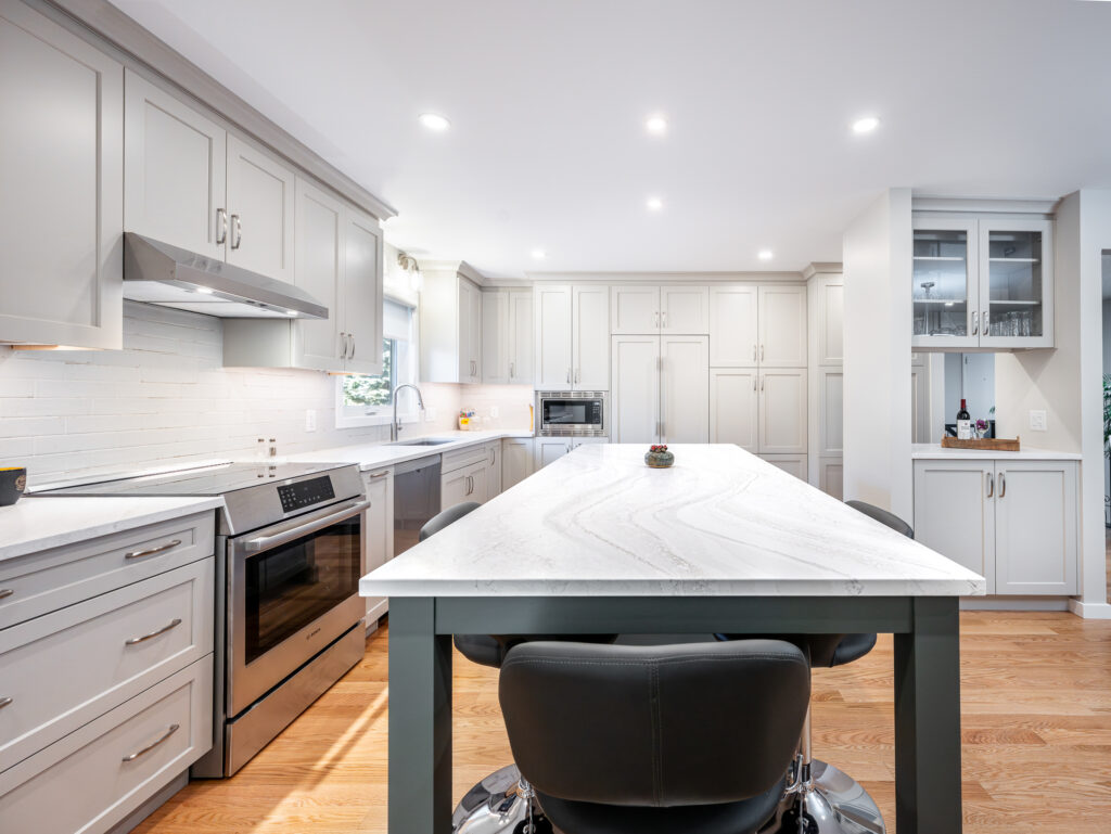 Modern kitchen with white cabinets, stainless steel appliances, marble island countertop, wooden floor, and recessed lighting. A black chair is at the island, and there’s a glass cabinet in the corner.