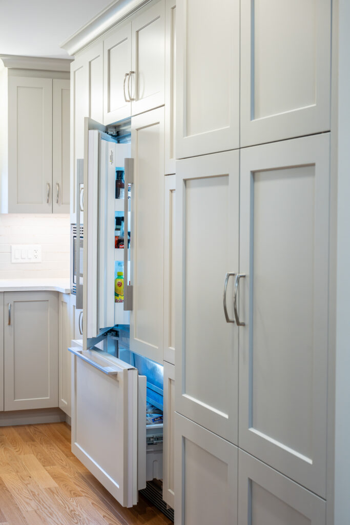 A modern kitchen with white cabinets. A built-in refrigerator with both the upper fridge and lower freezer doors open, revealing food items inside. The cabinets have silver handles and wood flooring is visible.