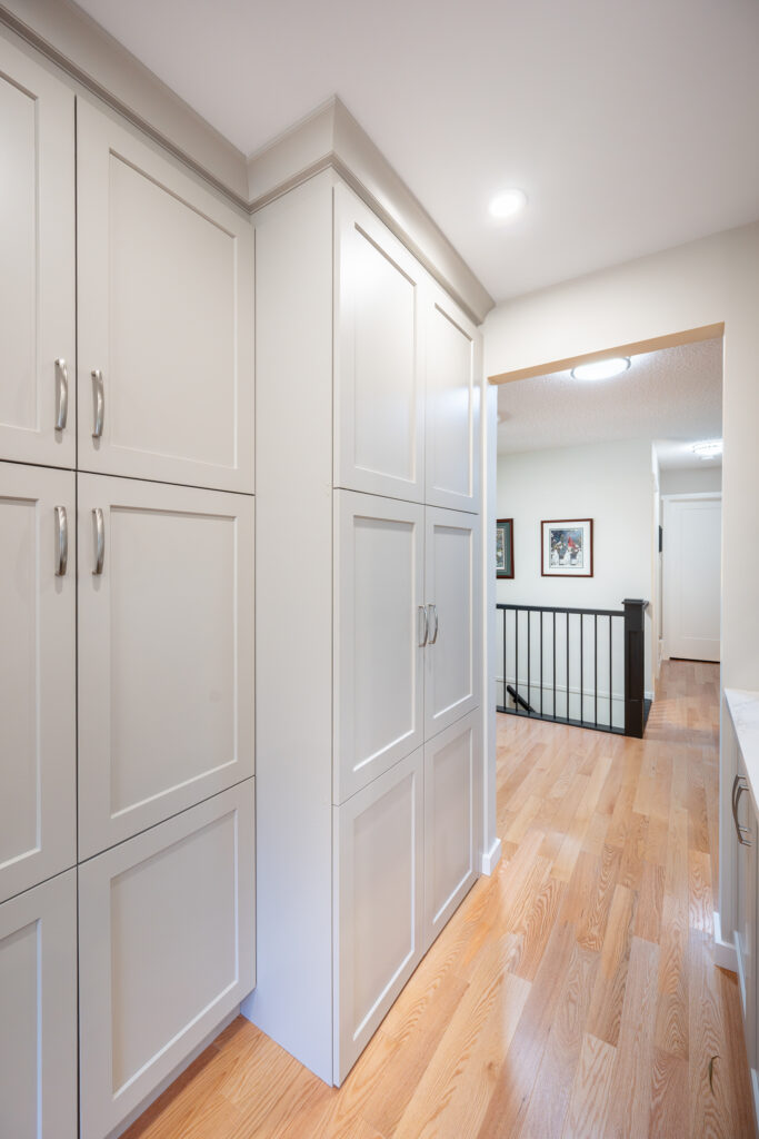 Bright hallway with light wood floors, white paneled cabinets on the left, a white ceiling with recessed lighting, and framed art on the walls near a black banister railing.