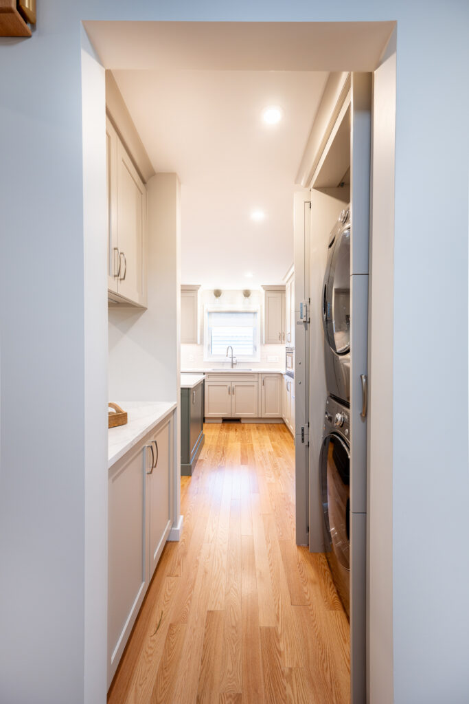 A narrow kitchen with light wood flooring, gray cabinets, and white countertops. A stacked washer and dryer unit is recessed into the right wall. Recessed lights illuminate the space, and a window is at the far end.