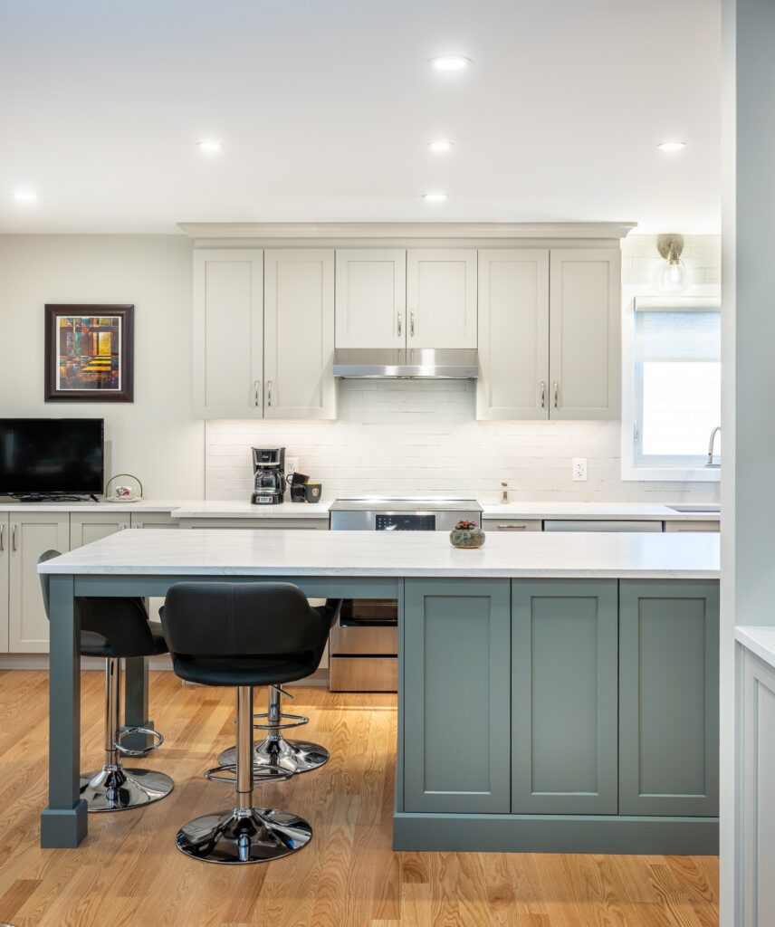 Modern kitchen with white cabinets, stainless steel appliances, a coffee maker, and a green island with two black chairs. Wood flooring, recessed lighting, a small TV, and a window create a bright, clean look.