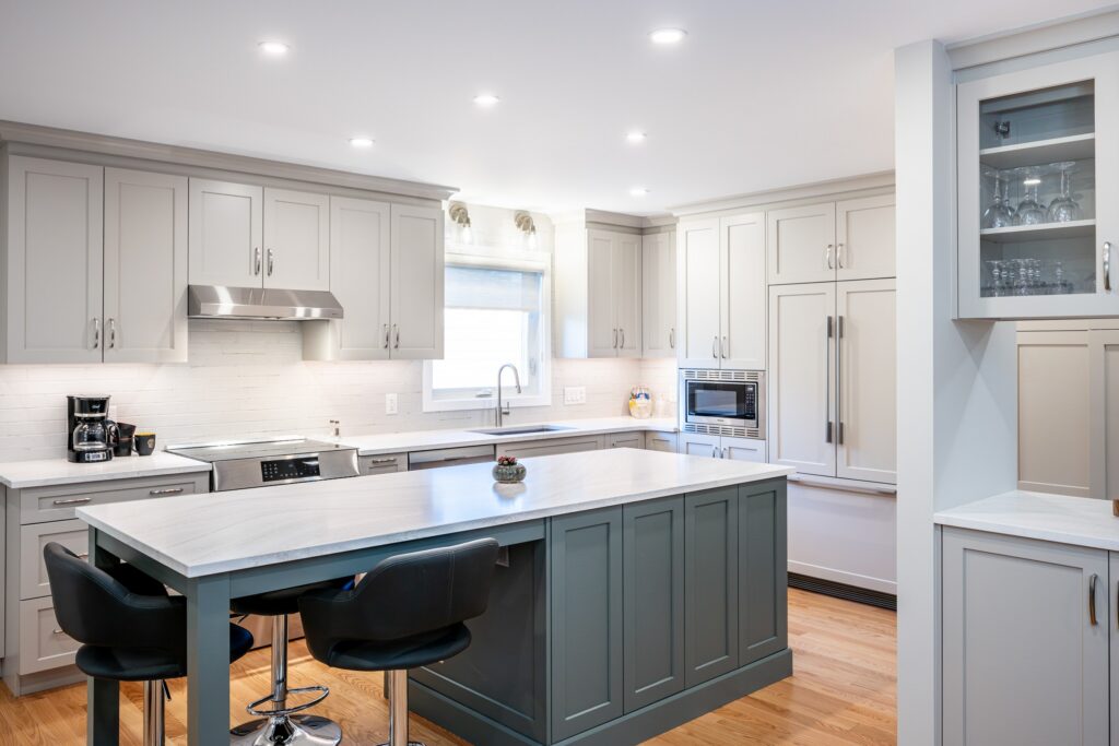 Modern kitchen with gray cabinets, stainless steel appliances, an island with a bowl of fruit, three black chairs, wood flooring, and bright recessed lighting.